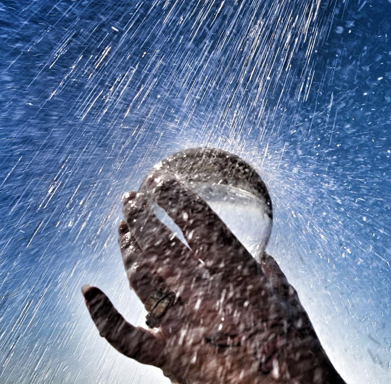 Image of a back lit hand holding a glass ball in front of a blue background. Water is falling on the objects and splashing, 