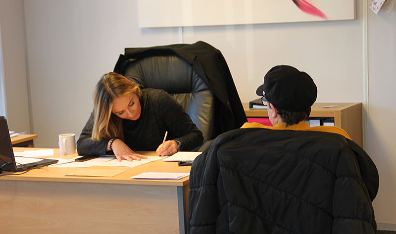 Photo of two people at desk. One woman is writing while the other watches.