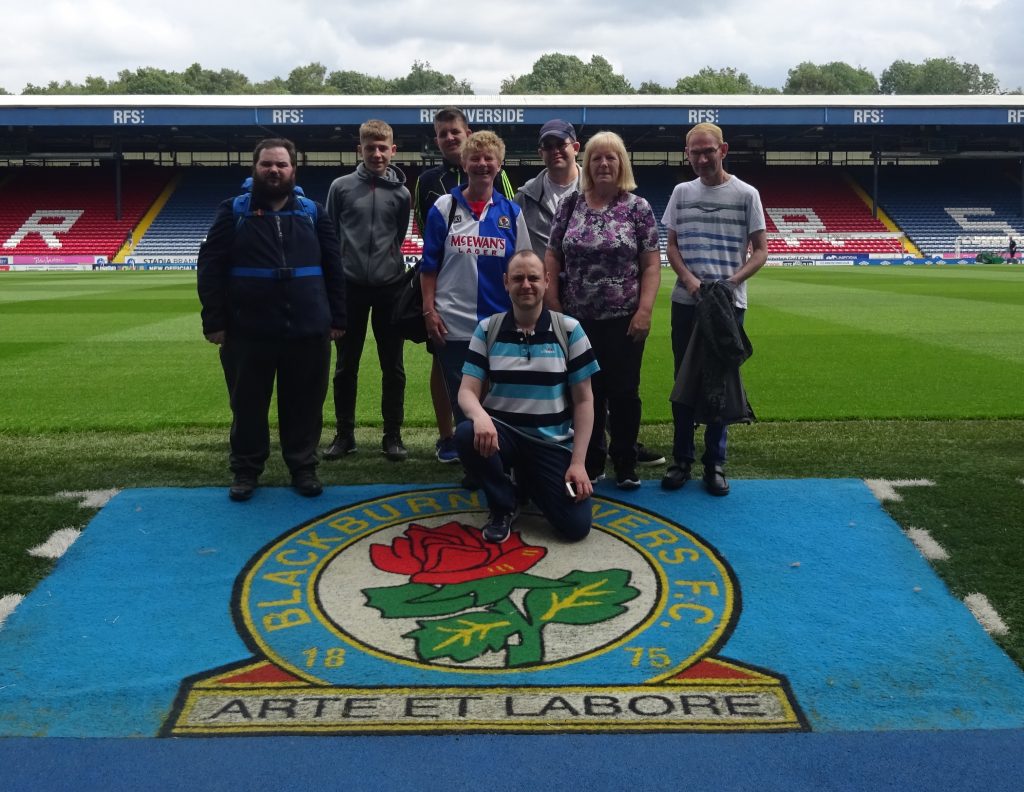 Group of 8 people in front of Blackburn Rovers football pitch on top of the club's logo.