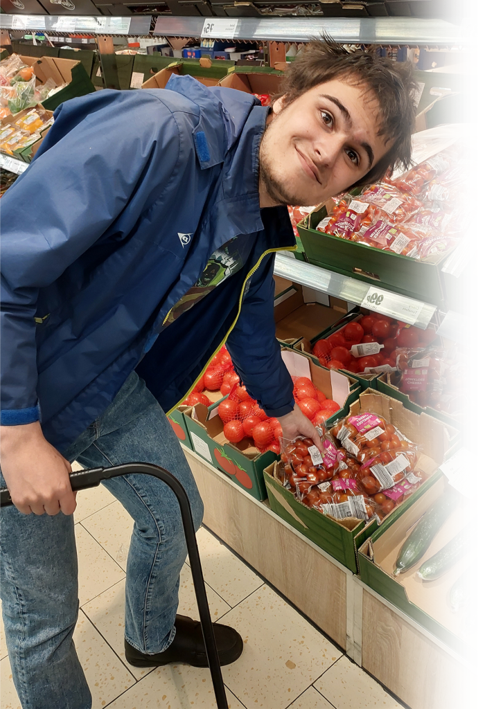 Photograph of a young man smiling in a supermarket, picking up fruit. 